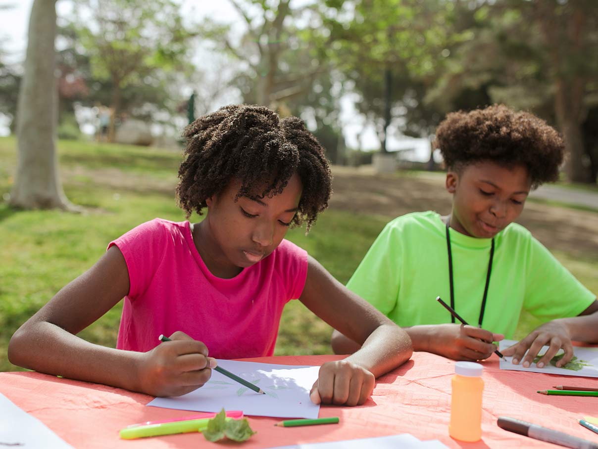 two children sat at a table outdoors, drawing toge