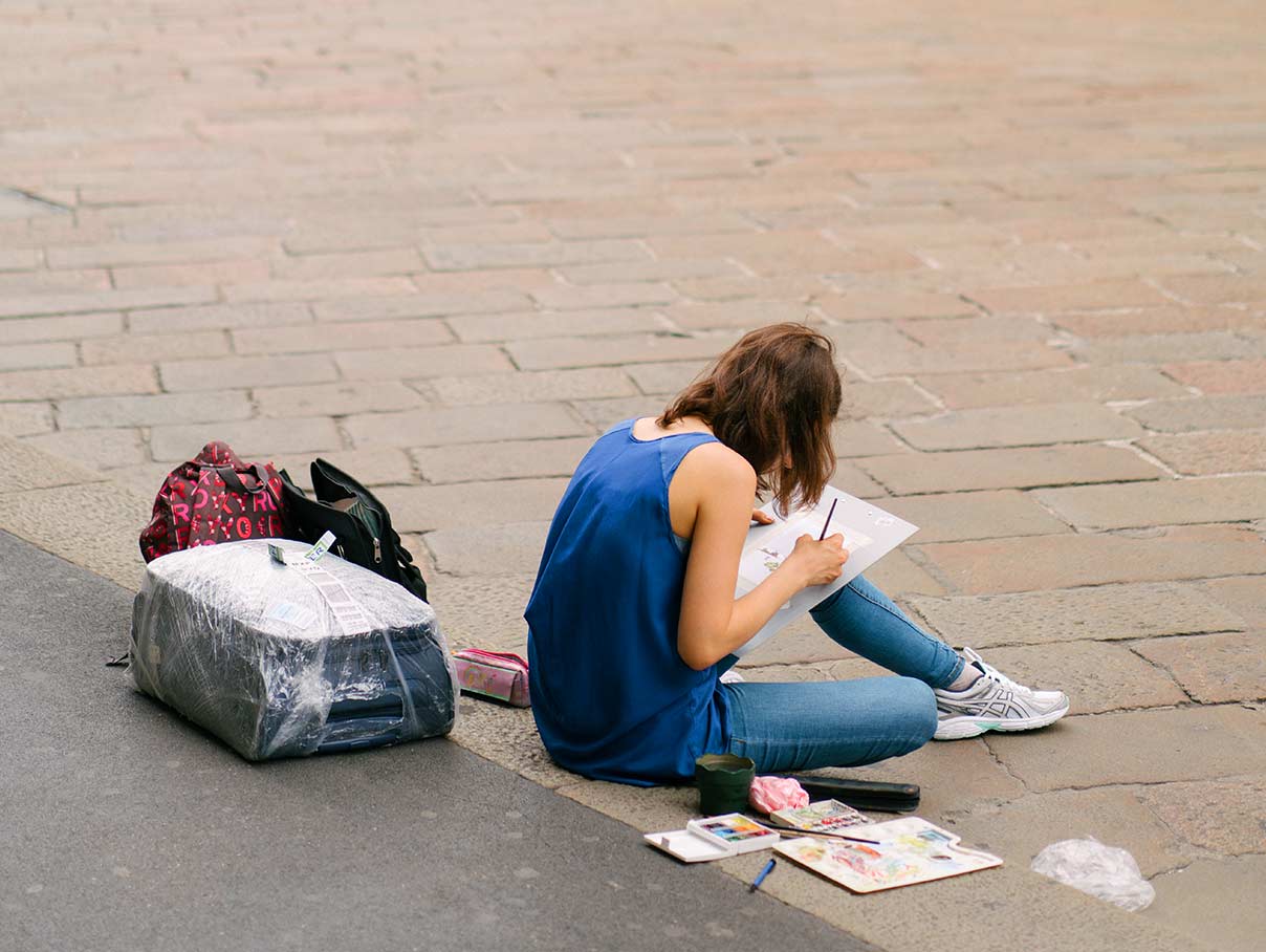 A woman sits on a piazza with her luggage, paintin
